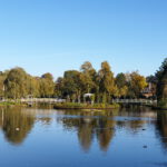 Im Vordergrund ist ein blauer, fast glatter See, auf dem ein paar Enten und Gänse schwimmen. In der Mitte liegt eine grün bewachsenen Insel, zu der von links und rechts je eine weiß angemalte Holzbrücke für Spazierende führt. Auf der Insel steht ein weißer Pavillion aus Metall mit Baldachin, dazu große und kleine Bäume. An den Bildränder sinlinks uns rechts sind ebenfalls Bäume, nämlich Birken und Weiden zu sehen. Sie alle spiegeln sich im Wasser des Sees. Oben im Bild ist der hellblaue, wolkenfreie Himmel.