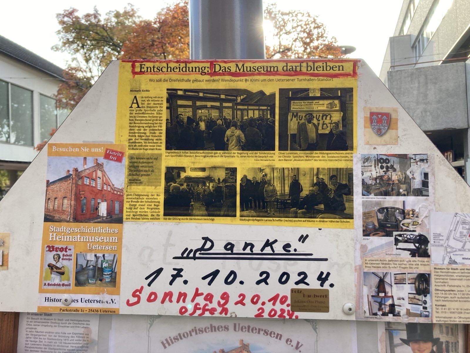 Das Foto zeigt ein Schild mit einer Collage. Sie Auf ihr sind Fotos des Gebäudes Gaswerkehaus in Uetersen zu sehen. Hier ist das Heimatmuseum untergebracht. Dazu gibt es einen Ausschnitte aus der Zeitung Uetersener Nachrichten. In dem Artikel wird über den Ortstermin berichtet, bei dem über den Standort der neuen 3-Feld-Halle beraten wurde. Der Titel des Artikels lässt vermuten, dass das Gebäude nicht abgerissen werden muss. Darunter ist noch ein Papier aufgeklebt mit den Worten "Danke - 17.10.2024" und "Sonntag, 20.10.2024 offen". Im Hintergrund sind die herbstlich gefärbten Blätter eines Baumes zu erkennen.