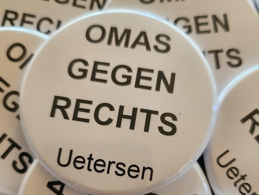 Das Foto zeigt den Ausschnitt eines Haufens von sechs Buttons zum Anstecken mit weißem Hintergrund und schwarzer Druck-Schrift. In der Mitte des Bildes ist ein Button komplett unverdeckt zu sehen. Auf ihm wie auf allen anderen steht in Großbuchstaben OMAS GEGEN RECHTS in drei Zeilen und darunter in Normalschrift Uetersen.