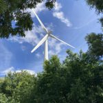 Vor blauem Himmel mit weißen Wölkchen ist in der Mitte des Fotos der obere Teile eines weißen Windrads zu sehen. in der unteren Hälfte des Fotos ist der untere Teil des Windrads von grünen Bäumen und Buschwerk verdeckt. Und er oberen linken Ecke des Fotos ragen Äste mit grünem Laub ins Bild.