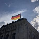 Das Foto zeit den oberen Teil eines der vier eckigen Türme des Reichstagsgebäudes in Berlin. Auf dem Dach des Turms flattert die Fahne der Bundesrepublik Deutschland im Wind vor einem sonnig blauen Himmel mit weißen Wolken.