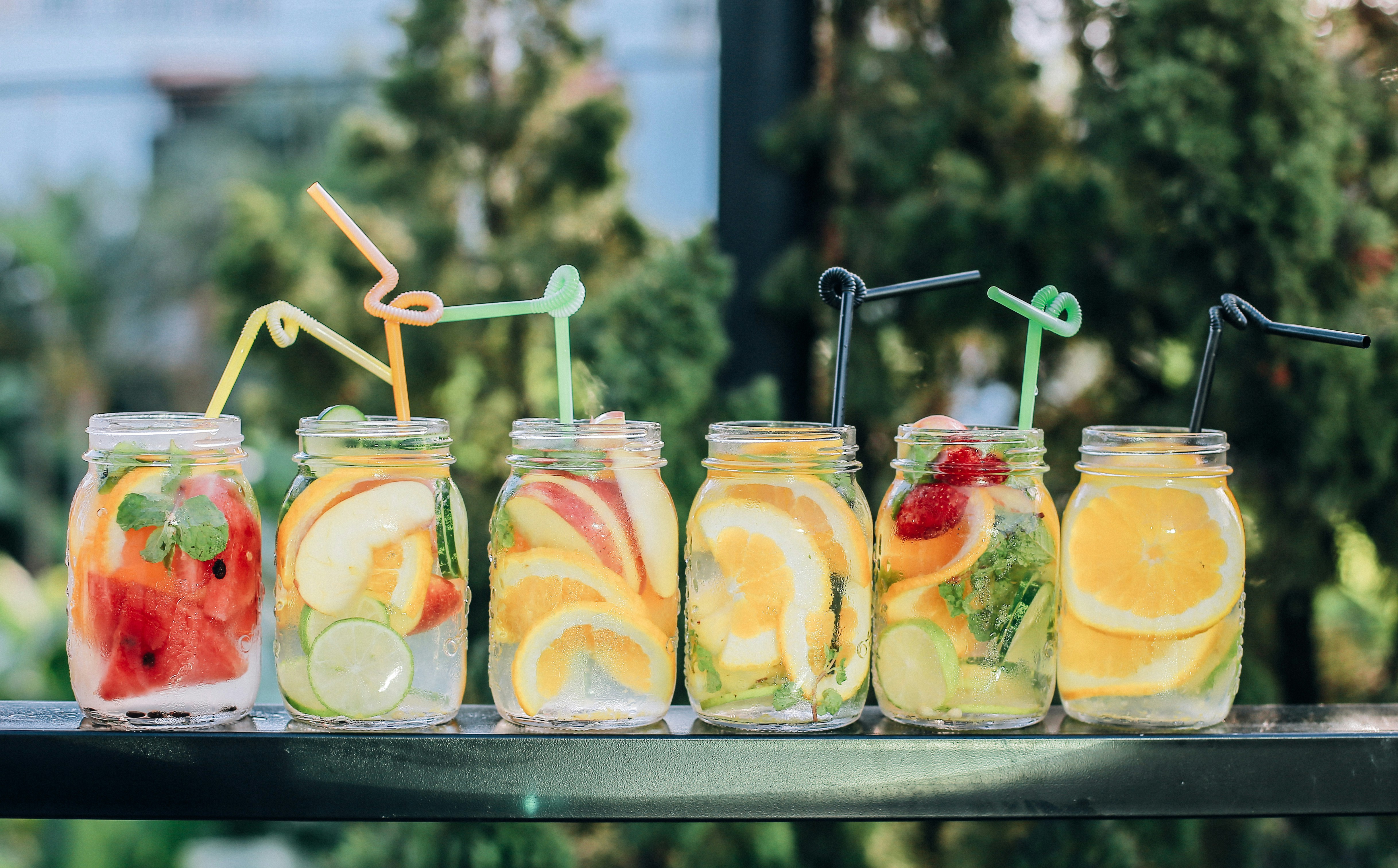 Das Foto zeigt einen Tisch von vorn. Auf ihm stehen 6 große Einweck-Gläser aus klarem Glas ohne Deckel, die mit frischem, geschnittenem Obst und Zitronemwasser gefüllt sind. Zu erkennen sind Spalten von Melone, Apfel, Zitrone, Orange, garniert mit Minze. In jedem Glas steckt ein bunter gedrehter Trinkhalm. Unscharf im Hintergrund sind grüne Büsche zu sehen wie in einem Garten.