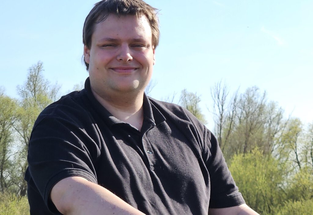 Das Foto zeigt einen jungen Mann in schwarzem Poloshirt. Er hat dunkelbraunes, kurzes Haar. Im Hintergrund sieht man grüne Bäume und Büsche, darüber den blauen Himmel.