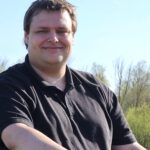 Das Foto zeigt einen jungen Mann in schwarzem Poloshirt. Er hat dunkelbraunes, kurzes Haar. Im Hintergrund sieht man grüne Bäume und Büsche, darüber den blauen Himmel.