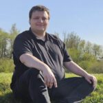 Das Foto zeigt einen jungen Mann in schwarzem Poloshirt und schwarzer Hose. Er hat dunkelbraunes, kurzes Haar und sitzt auf einem Deich auf einer grünen Wiese. Im Hintergrund sieht man grüne Bäume und Büsche, darüber den blauen Himmel.