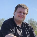 Das Foto zeigt einen jungen Mann in schwarzem Poloshirt. Er hat dunkelbraunes, kurzes Haar. Im Hintergrund sieht man grüne Bäume und Büsche, darüber den blauen Himmel.