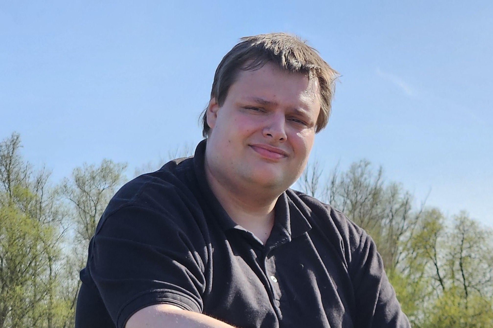 Das Foto zeigt einen jungen Mann in schwarzem Poloshirt. Er hat dunkelbraunes, kurzes Haar. Im Hintergrund sieht man grüne Bäume und Büsche, darüber den blauen Himmel.