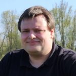 Das Foto zeigt einen jungen Mann in schwarzem Poloshirt. Er hat dunkelbraunes, kurzes Haar. Im Hintergrund sieht man grüne Bäume und Büsche, darüber den blauen Himmel.