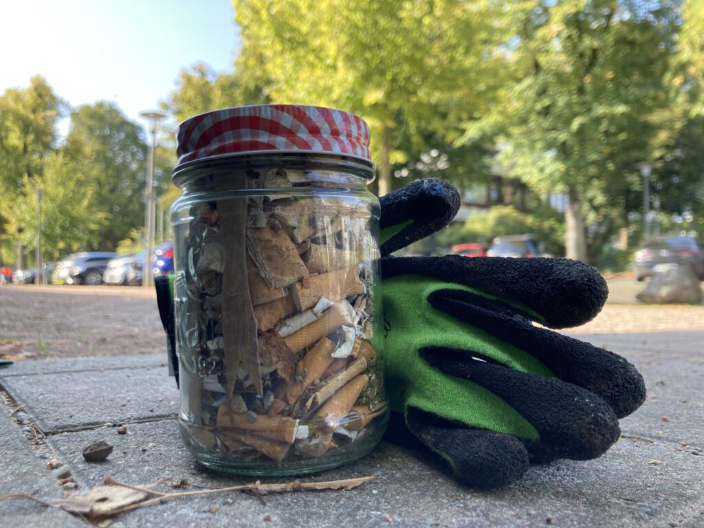 In der Mitte des Fotos im Vordergrund groß ist ein Marmeladeglas mit Schraubverschluss zu sehen, gefüllt mit aufgesammelten Zigarettenkippen, daneben ein paar Handschuhe für die Gartenarbeit. Beides steht auf dem Fußweg. Die Straße mit parkenden Autos und grünen Bäumen ist verschwommen im Hintergrund zu erkennen. Oben links im Bild sieht man ein Stück blauweißen Himmel.