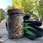 In der Mitte des Fotos im Vordergrund groß ist ein Marmeladeglas mit Schraubverschluss zu sehen, gefüllt mit aufgesammelten Zigarettenkippen, daneben ein paar Handschuhe für die Gartenarbeit. Beides steht auf dem Fußweg. Die Straße mit parkenden Autos und grünen Bäumen ist verschwommen im Hintergrund zu erkennen. Oben links im Bild sieht man ein Stück blauweißen Himmel.