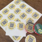 Das Foto zeig eine Holztischplatte von oben, au der ein Blatt Papier mit 24 Sonnenblumen mit QRCode und er Mitte. Vier davon sind bereits ausgeschnitten. Ein rund ausgeschnittener Sonnenblumen QRCode liegt in einem geöffneten roten Taschenaschenbecher. Auf dem rechts daneben liegenden Deckel steht "sollte, hätte, könnte müsste" jeweils rot durchgestrichen und "MACHEN!" in einer gelben Sonnenblume, alles auf grünem Grund. Die Schere liegt oben links im Bild.