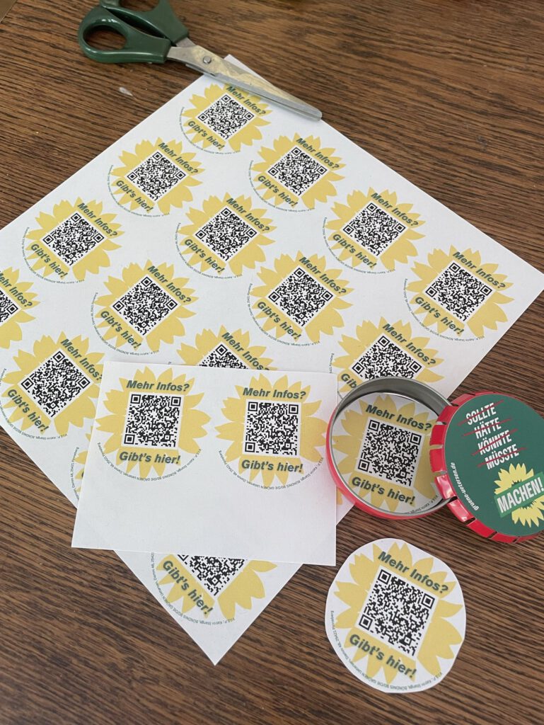 Das Foto zeig eine Holztischplatte von oben, au der ein Blatt Papier mit 24 Sonnenblumen mit QRCode und er Mitte. Vier davon sind bereits ausgeschnitten. Ein rund ausgeschnittener Sonnenblumen QRCode liegt in einem geöffneten roten Taschenaschenbecher. Auf dem rechts daneben liegenden Deckel steht "sollte, hätte, könnte müsste" jeweils rot durchgestrichen und "MACHEN!" in einer gelben Sonnenblume, alles auf grünem Grund. Die Schere liegt oben links im Bild.