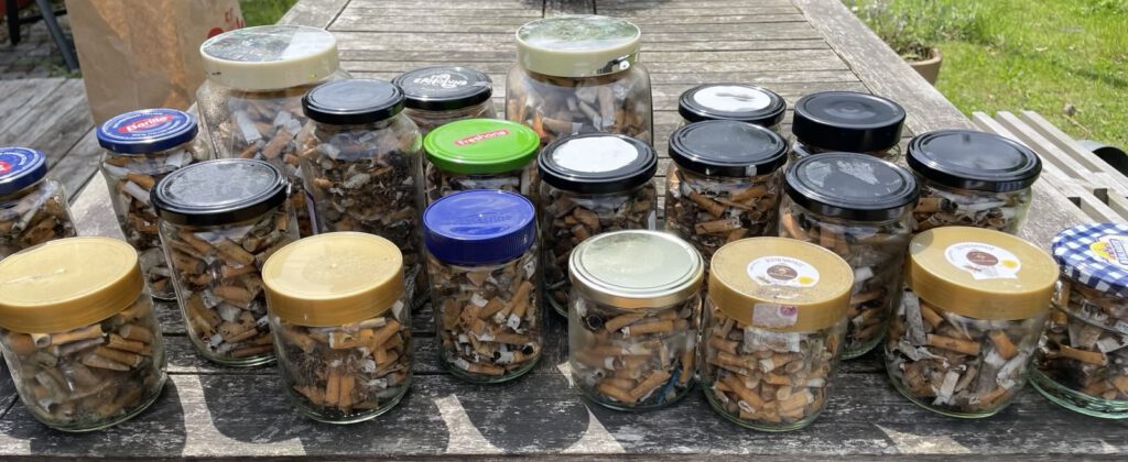 Das Foto zeigt einen Gartentisch aus Holz von schräg oben. Auf dem Tisch im Vordergrund stehen 21 unterschiedlich große Schraubverschlussgläser, alle randvoll gefüllt mit Zigarettenkippen.