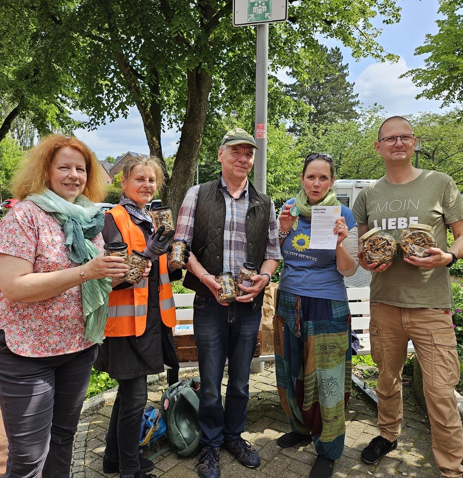 Das Foto zeigt eine Gruppe von fünf Menschen. Sie stehen neben einander und sehen alle freundlich bestimmt und ie Kamera. In den Händen halten sie Schraubverschlussgläser, gefüllt mit den gesammelten Zigarettenkippen. Im Hintergrund sieht man grüne Bäume, Büsche und den blauen Himmel mit ein paar weißen Wolken.