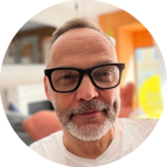 Das Bild zeigt das Gesicht eines Mannes mit kurzen graublonden Haaren und mit grauweißem gestutzten Bart und schwarzer Brille. Er trägt ein weißes T-Shirt. Das ist Dirk Neubauer. Er lächelt verschmitzt.