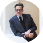 Das Foto zeigt einen Mann im dunkelgrauen Anzug mit Pullunder und Krawatte und hellgrauem Hemd. Er trägt eine schwarze Brille und lehnt mit seinem rechten Arm auf einer weißen Balustrade. Das ist Konstantin von Notz. Er lächelt verschmitzt.