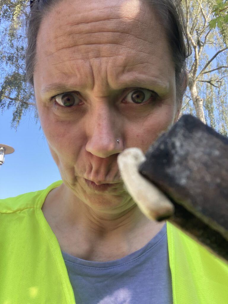 Das Foto zeigt das Porträt einer Frau, die die Stirn kraus und den Mund verzieht. Im Vordergrund sieht man die Spitze einer Zange, die eine einzelne Zigarettenkippe in die Kamera hält. Die Frau trägt eine neongelbe West über einem jeansblauen Shirt. Ihre Haare sind aus dem Gesicht gekämmt. Im Hintergrund sieht man blauen Himmel und die Blätter und Zweige eines Baumes.