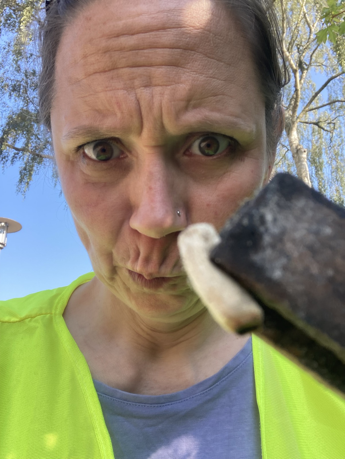 Das Foto zeigt das Porträt einer Frau, die die Stirn kraus und den Mund verzieht. Im Vordergrund sieht man die Spitze einer Zange, die eine einzelne Zigarettenkippe in die Kamera hält. Die Frau trägt eine neongelbe West über einem jeansblauen Shirt. Ihre Haare sind aus dem Gesicht gekämmt. Im Hintergrund sieht man blauen Himmel und die Blätter und Zweige eines Baumes.