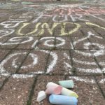 Das Bild zeigt aus der Forschperspektive die Pflasterung eines Platzes. Im Vordergrund liegt Malkreise in Blau, Rot, Gelb, Grün und Weiß auf dem Pflaster, das mit dieser Kreis bemalt und beschrieben wurde. Im Hintergrund sind unscharf drei Menschen zu erkennen, die gerade auf die steine malen.