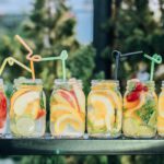 Das Foto zeigt einen Tisch von vorn. Auf ihm stehen 6 große Einweck-Gläser aus klarem Glas ohne Deckel, die mit frischem, geschnittenem Obst und Zitronemwasser gefüllt sind. Zu erkennen sind Spalten von Melone, Apfel, Zitrone, Orange, garniert mit Minze. In jedem Glas steckt ein bunter gedrehter Trinkhalm. Unscharf im Hintergrund sind grüne Büsche zu sehen wie in einem Garten.